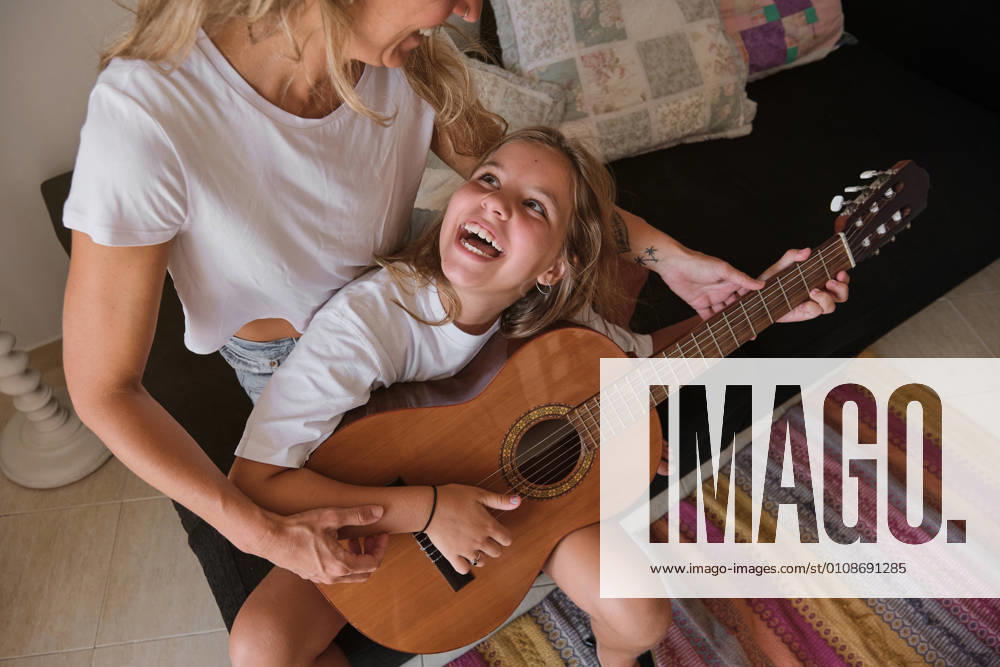 IMAGO Creative: Bildung, Musikerziehung Blonde woman teaching a girl to play the guitar while looking at each other laughing sitting on a couch in a house, Model released
