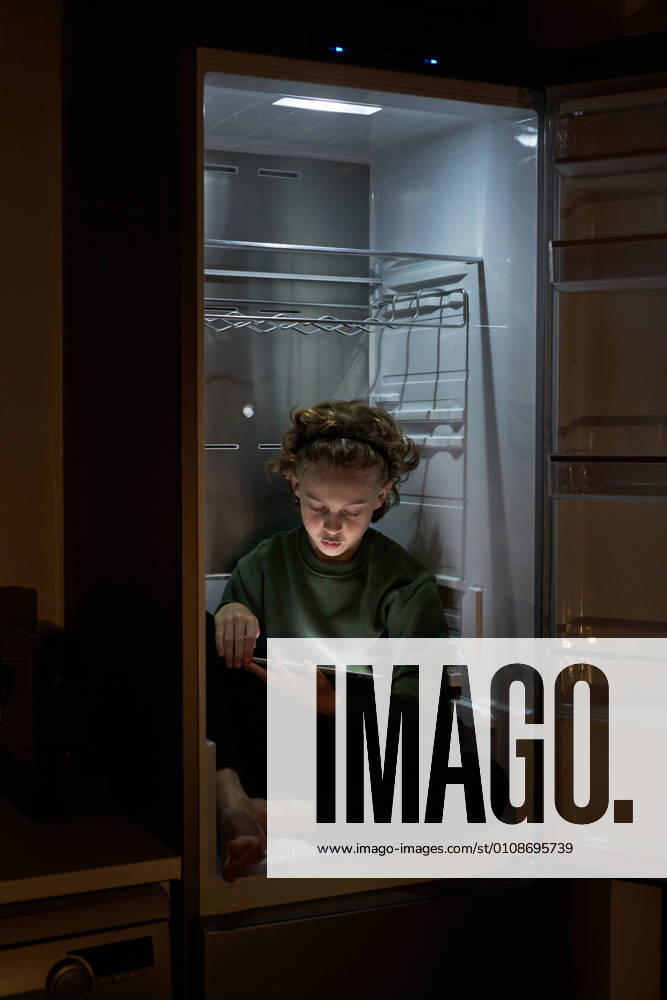 Calm relaxed kid sitting in empty fridge and using digital tablet at hot night