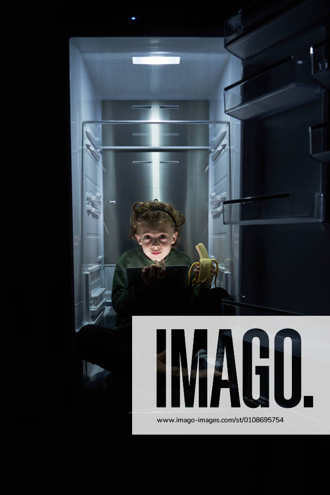Calm relaxed kid sitting in empty fridge with ripe banana and using digital tablet at hot night
