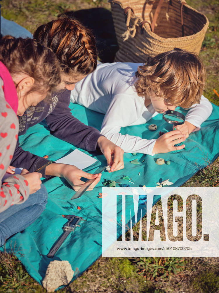 IMAGO Creative: N?chste Generation, Sozial- und klimabewusst Young lady with pencil writing in notebook and children gauging by ruler little plastic things and lying on coverlet on ground