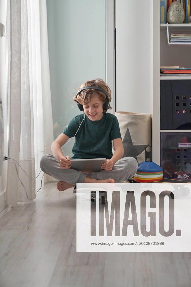 Cheerful preteen boy in headphones and casual clothes focusing on screen and smiling while sitting on skateboard and using digital tablet with pleasure in light modern room