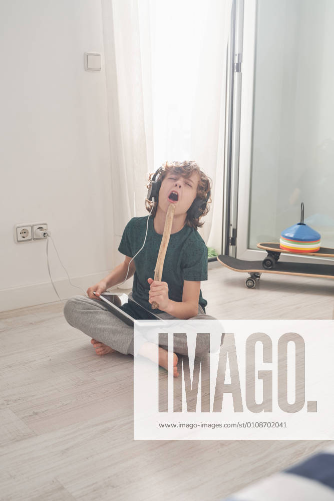 Cheerful preteen boy in headphones and casual clothes singing with closed eyes while using digital tablet in light modern room