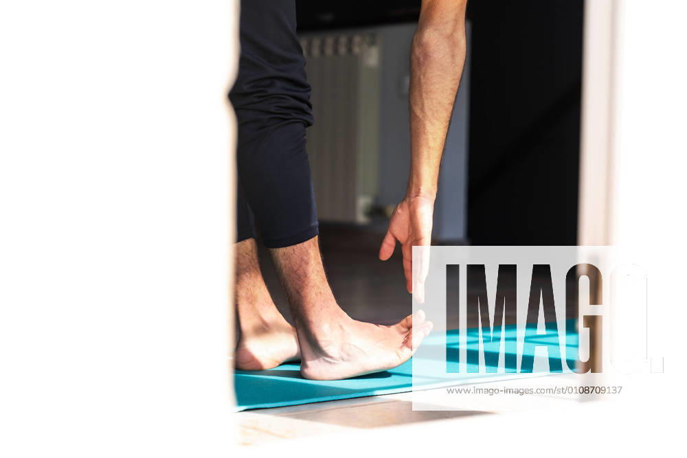 Side view of unrecognizable flexible barefoot man in sportswear doing standing forward bend exercise while training alone on sports mat at home
