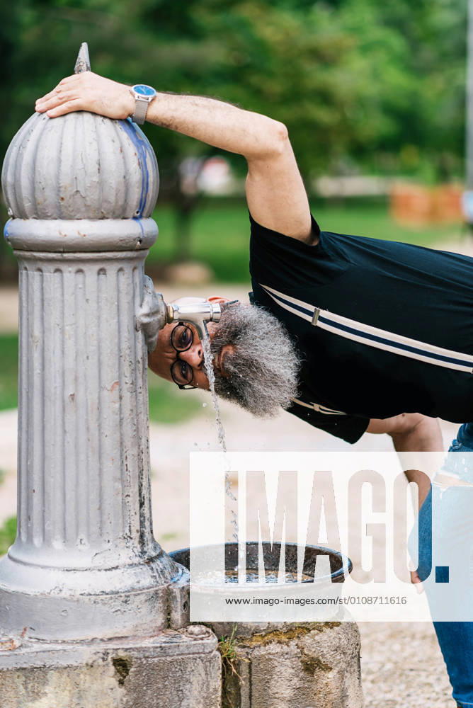 IMAGO Creative, Jahreszeit Sommer, Sommer in der Stadt Content old male hipster in trendy clothes drinking water from fountain in city while looking at camera, Model released