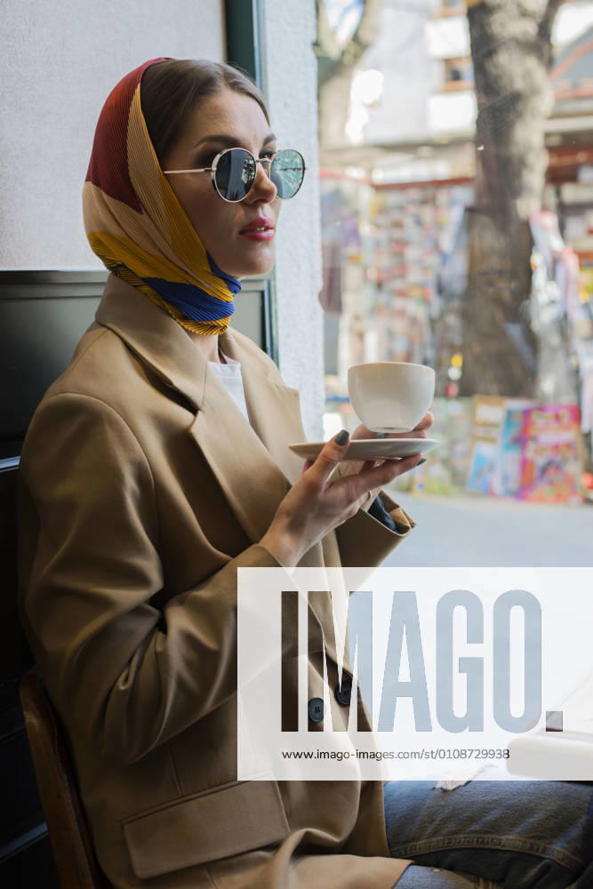Side view of trendy young female in eyeglasses and shawl on head enjoying coffee while sitting with crossed legs in cafeteria near window in afternoon