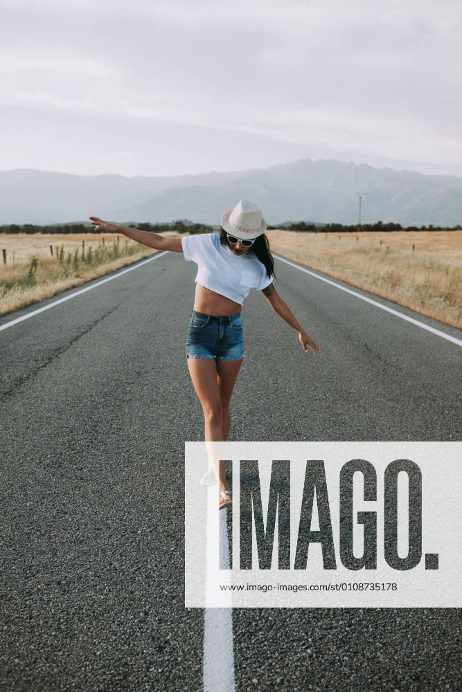 Serene female traveler in summer wear walking with outstretched arms along empty asphalt roadway on background of mountainous landscape and enjoying freedom