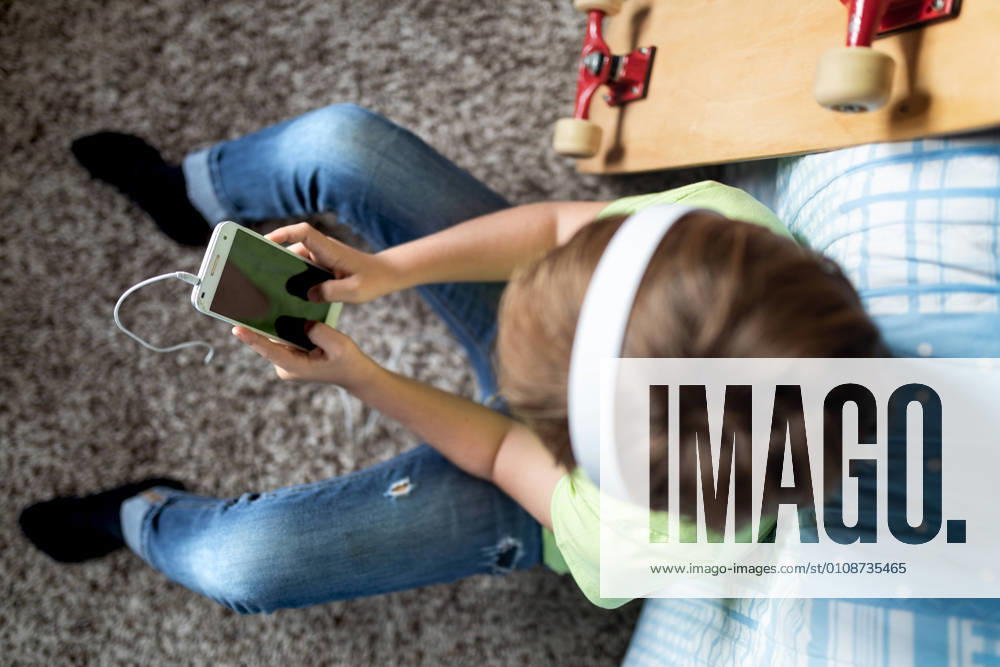 Little boy with headphones listening to music and chatting with friends in social network while sitting near skateboard in bedroom
