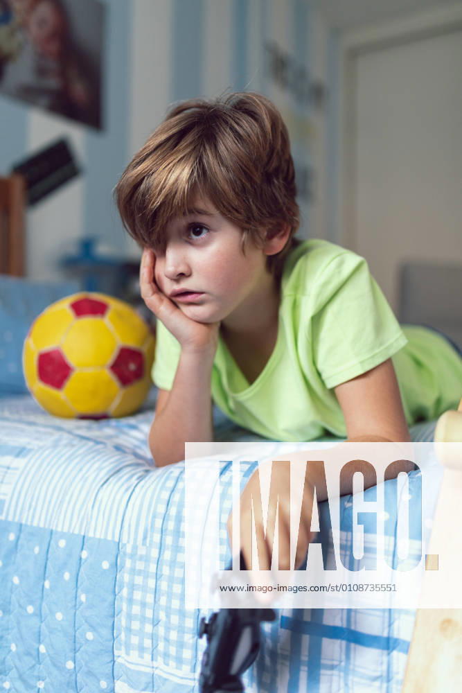 Bored little boy in casual clothes lying on bed near ball and skateboard unhappy with self isolation at home