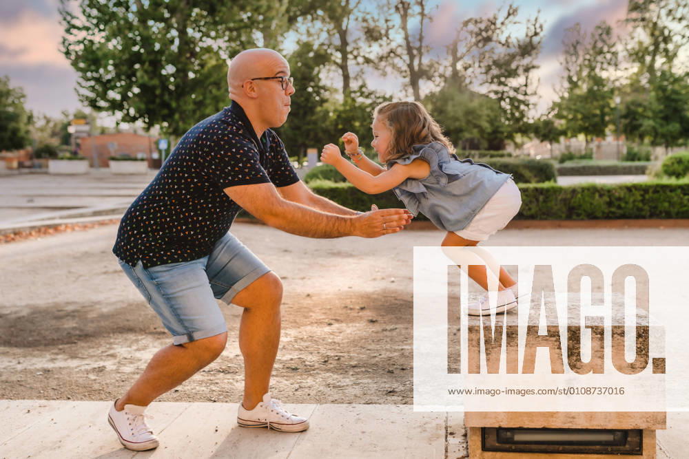 IMAGO Creative: Sommer, Sommertage im Stadtpark Side view of father standing in park and ready for catching playful daughter jumping from stone parapet while entertaining together in city, Model released