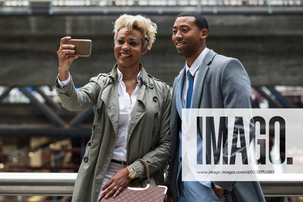 Elegant trendy black coworkers laughing happily while taking selfie with smartphone on station., Model released