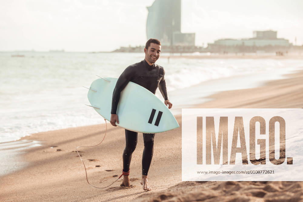 IMAGO Creative, Jahreszeit Sommer, Sommer in der Stadt Happy adult man in wetsuit carrying surfboard walking on sandy beach in sunlight, Barcelona