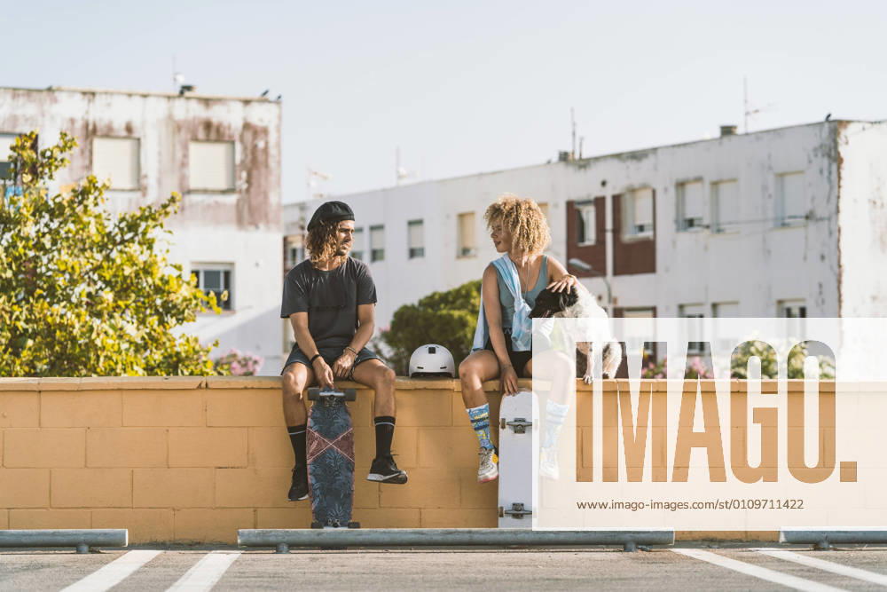 IMAGO Creative, Urban Lifestyle IMAGO Creative, Jahreszeit Sommer, Sommer in der Stadt Friends with skateboard and dog talking while sitting on retaining wall in city model released Symbolfoto DAMF00555