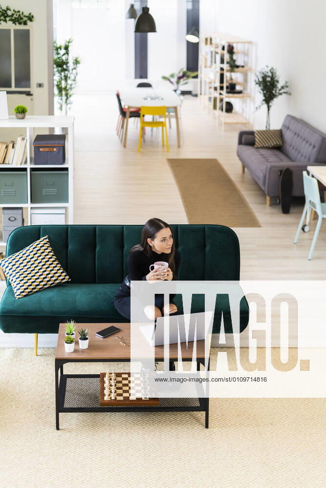 Young woman holding coffee cup while looking away in living room at home model released Symbolfoto property released GIOF09414