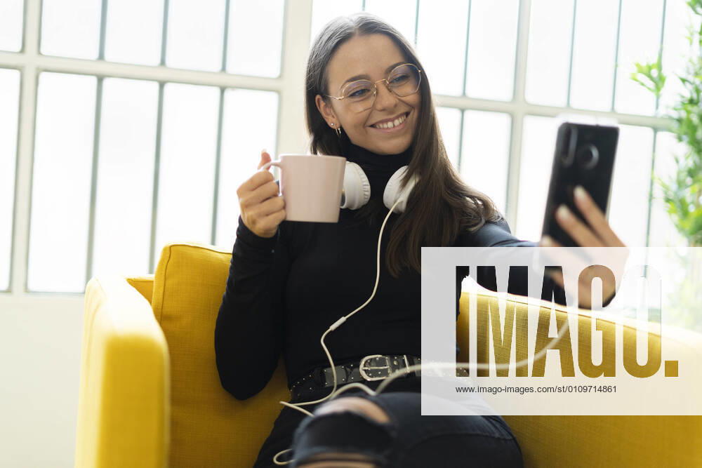 Smiling young female blogger taking selfie with coffee cup and headphones against window at home model released Symbolfoto property released GIOF09444
