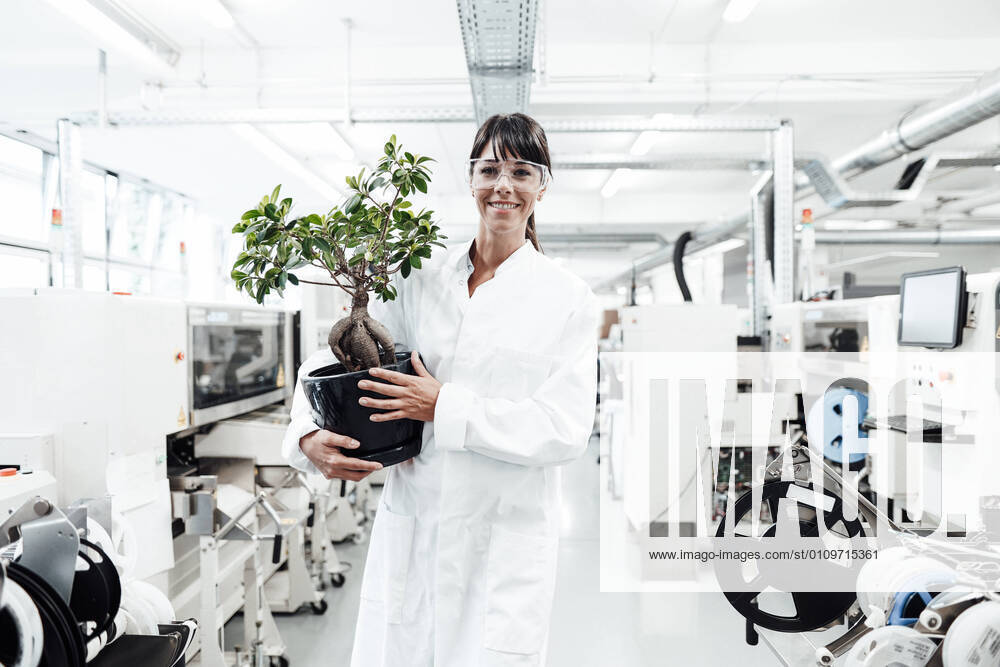 IMAGO Creative: Klimawandel - Zukunft und Innovation Smiling female scientist holding potted plant while standing amidst machineries at laboratory model released Symbolfoto property released JOSEF02177