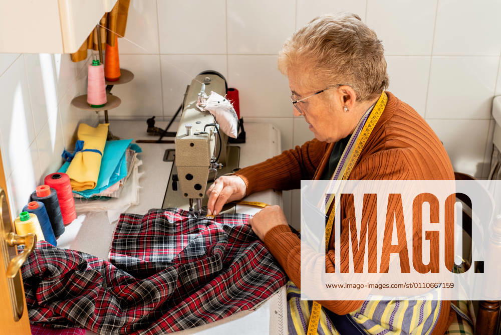 Side view of concentrated senior seamstress sitting at sewing machine and stitching piece of cloth while working in workshop, Model released, Property released