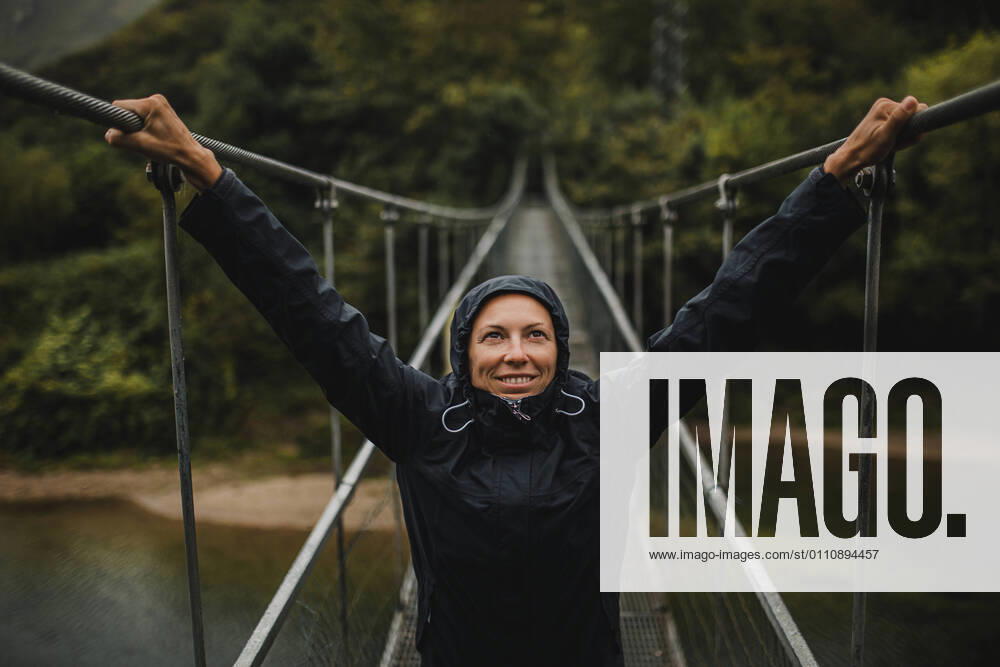 IMAGO Creative: Ende des Sommers Smiling woman wearing raincoat with arms raised standing on suspension bridge over Sella river in forest model released Symbolfoto DMGF00421