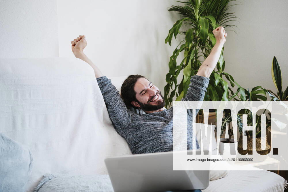 Young man with laptop stretching hand while sitting on sofa at home model released Symbolfoto property released EBBF01808