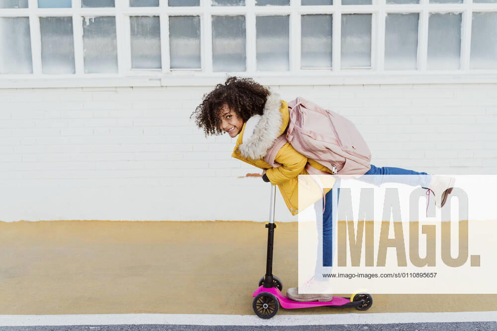 Portrait of cheerful girl with curly hair riding push scooter on road by building model released Symbolfoto EBBF01968