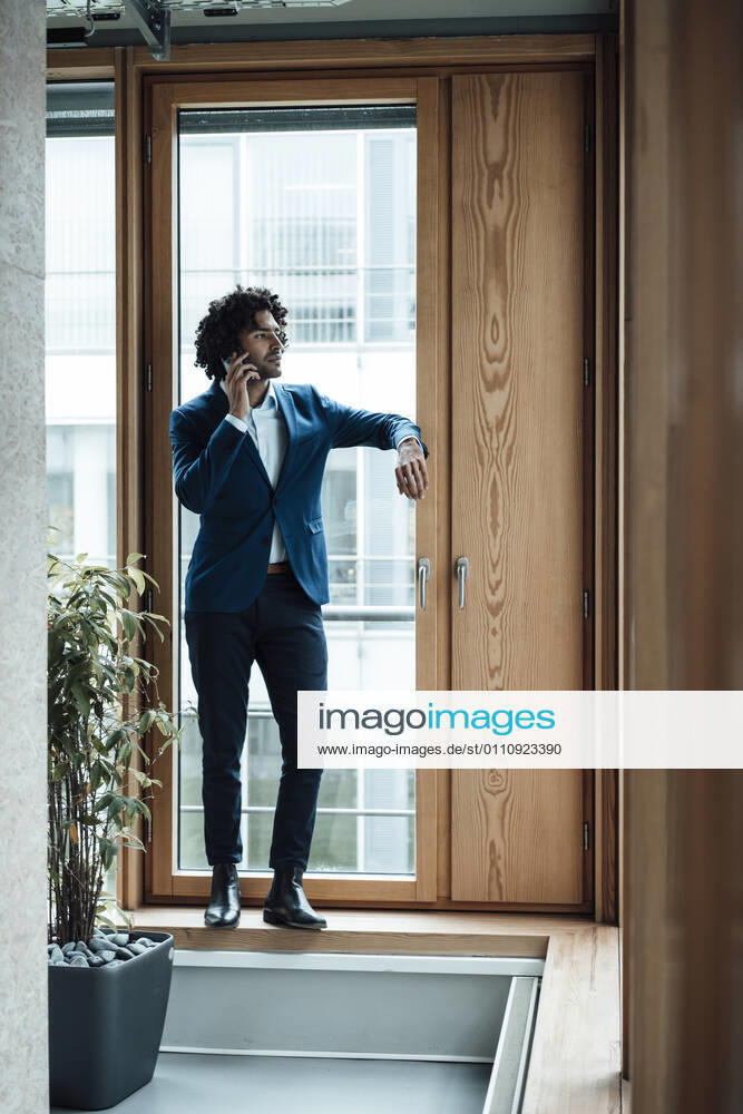 Male entrepreneur talking on mobile phone while standing against window at office model released Symbolfoto property released JOSEF02853