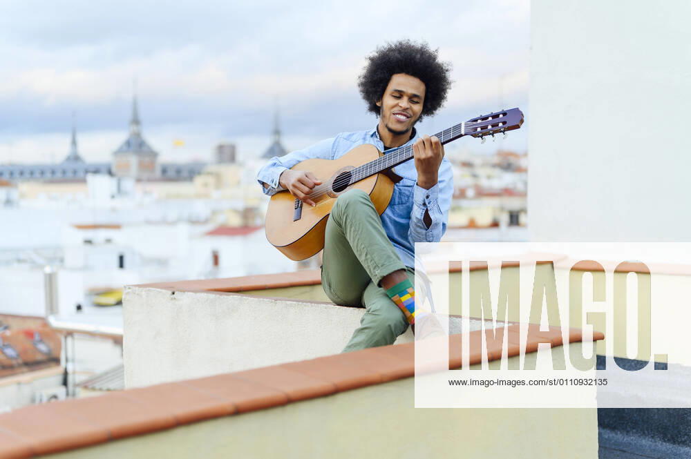 IMAGO Creative: Bildung, Musikerziehung Tag der Musik am 21. Juni Young man with curly hair playing acoustic guitar while sitting on terrace model released Symbolfoto PGF00320