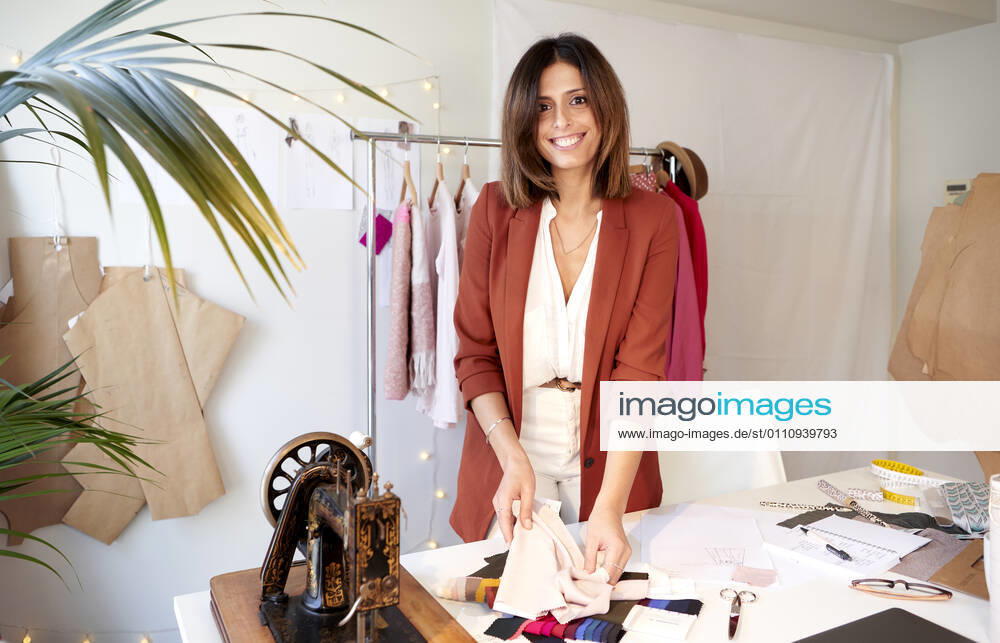 Smiling female design professional holding fabric swatch at desk against clothes rack in atelier model released Symbolfoto property released VEGF03413