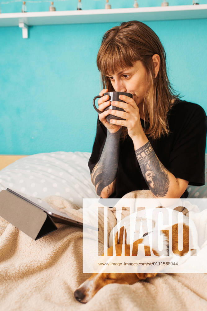 Positive young female in black shirt enjoying hot tea and watching video on tablet while resting on comfortable bed with adorable friendly dog
