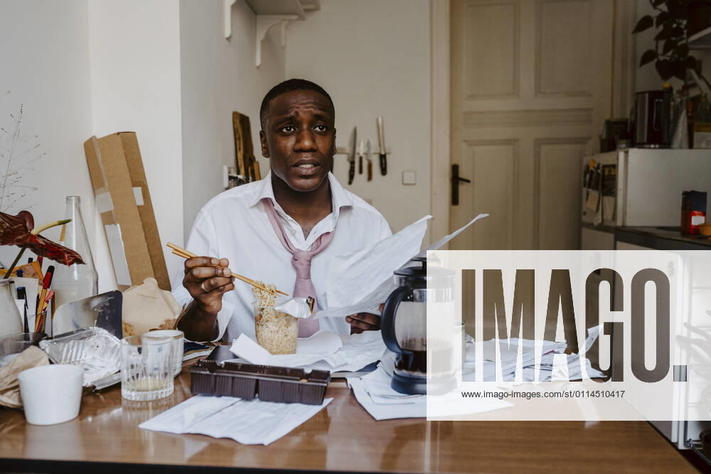 Thoughtful male entrepreneur with paper document having noodles at table in living room Germany, Berlin, model released, property released