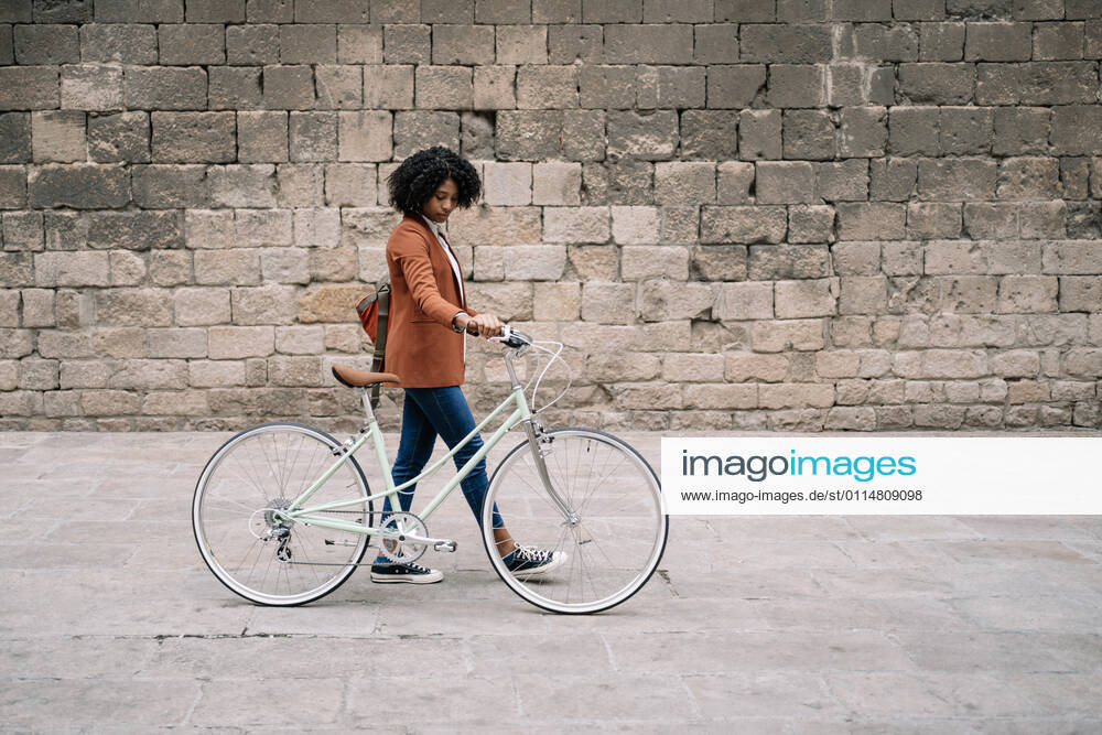 Side view of black woman in smart casual style walking along street with bike and looking down