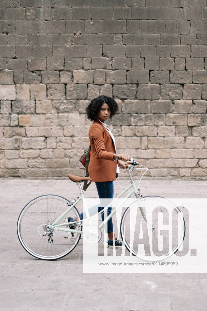 Side view of black woman in smart casual style walking along street with bike and looking at camera