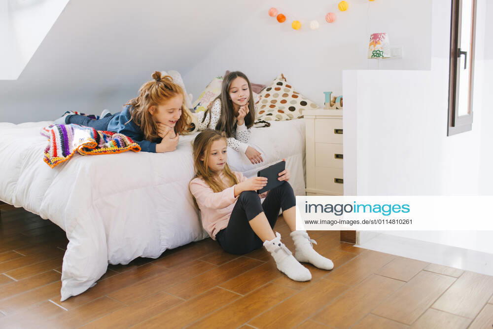 Cute little girls playing games on tablet and resting on soft bed while spending weekend together