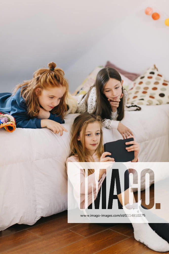 Cute little girls playing games on tablet and resting on soft bed while spending weekend together