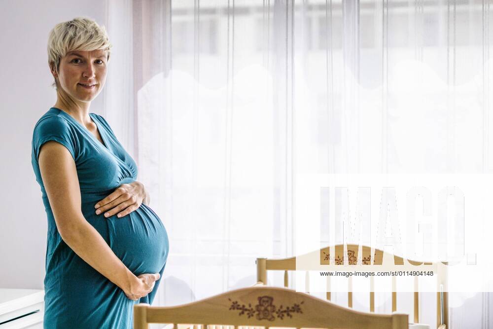 Pregnant woman holding her belly by baby bed next to window