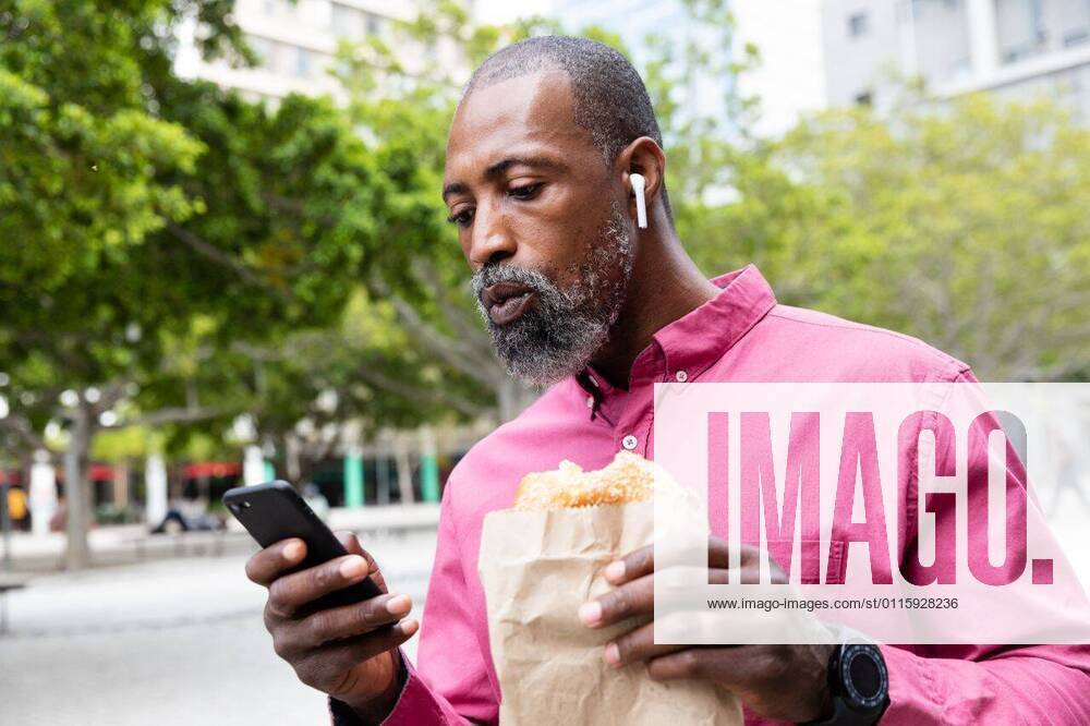 African American man using his phone and eating a takeaway sandwich African American man out and about in the city streets during the day, wearing pink shirt and earphones, having a takeaway snack and using a smartphone., model released, , property released, Cape Town, South Africa, 11.03.2020, 1334722.jpg, people,real people,commuting,commuter,travel,city,creative,individuality,lifestyle,street fashion,modern,on the go,digital nomad,independent,casual,street,smartphone,mobile phone,technology,phone,holding,using,communication,connection,interacting,earphones,listening,musing,smartwatch,takeaway,to go,snack,lunch,street food,eating,paper bag Mature Adult,Man,Male,Black Outdoors,Side view,Day,Half length