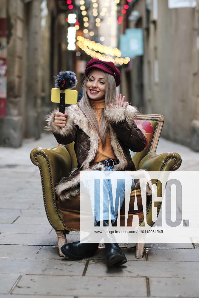 Smiling woman in fur coat vlogging through smart phone while sitting on armchair at alley model released Symbolfoto AGOF00083