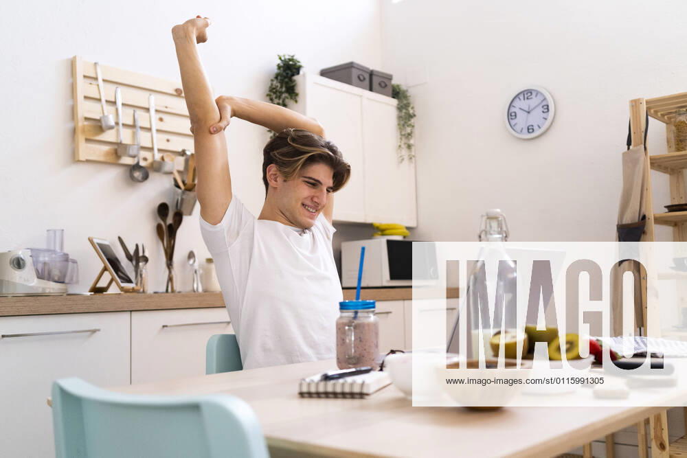 Young man stretching his hand while sitting at table with laptop in kitchen model released Symbolfoto property released GIOF11802