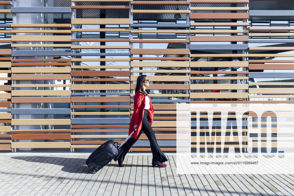 Businesswoman walking on footpath with suitcase by wooden wall during sunny day model released Symbolfoto JRVF00340