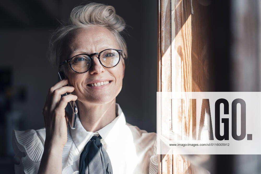 Smiling businesswoman with eyeglasses looking away while talking on smart phone at window in home office model released Symbolfoto property released MOEF03651