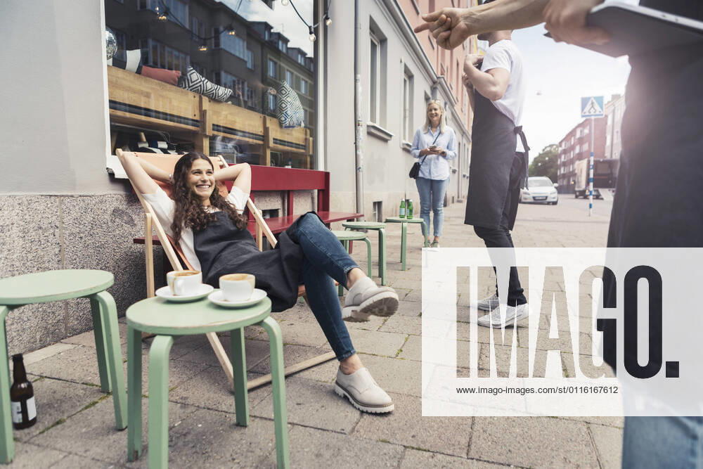 Smiling female owner with hands behind head sitting on deck chair outside coffee shop Sweden, Malmo, model released, property released