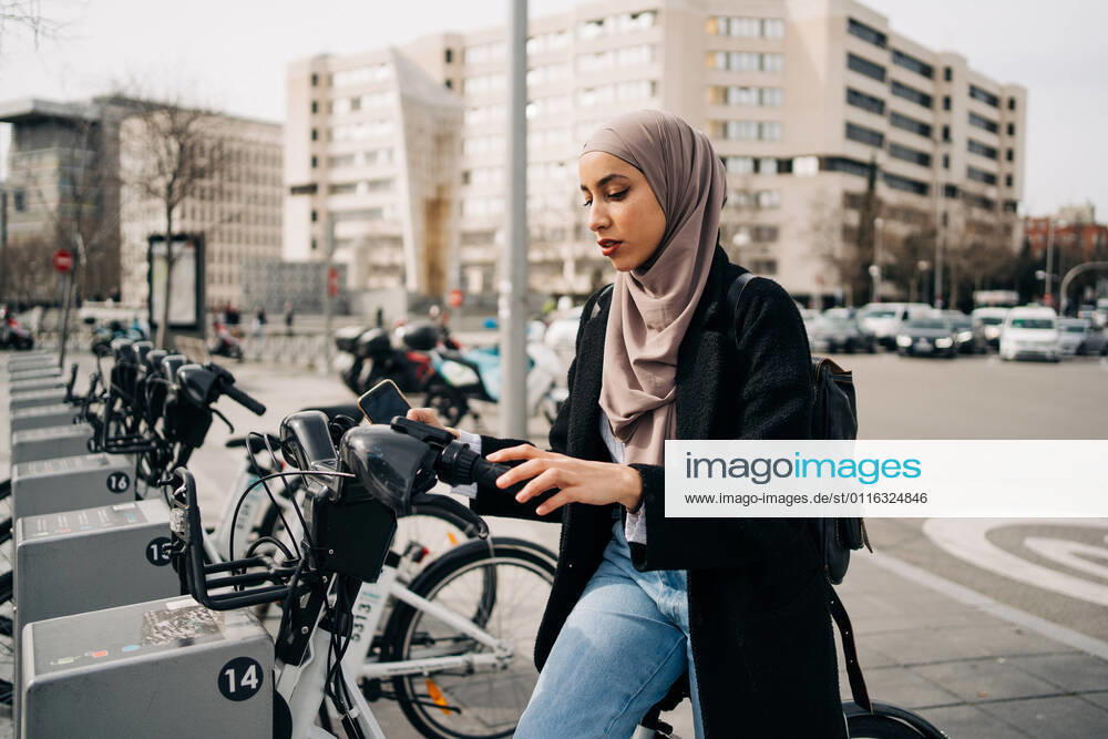 Side view of Muslim female in headscarf using bicycle sharing system in city