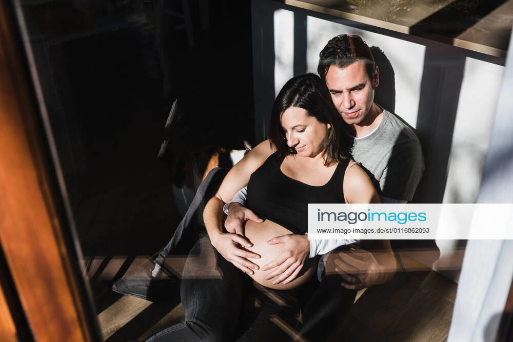 Through window from above of tranquil man hugging pregnant woman from behind and touching belly while sitting together on floor in room lit by sunlight, Model released