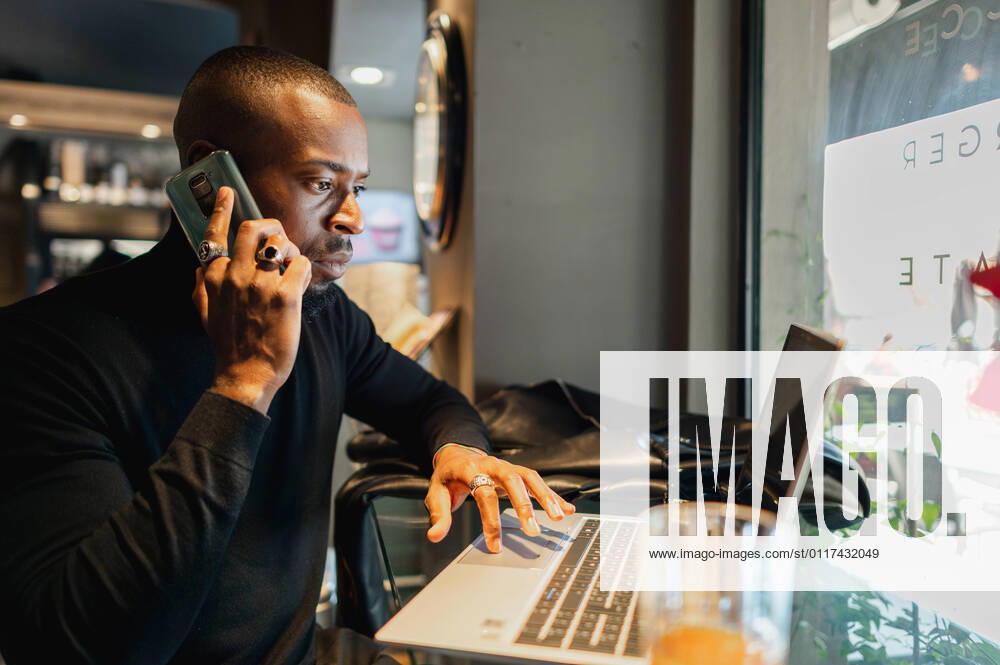 Side view of focused African American male entrepreneur working remotely in cafe while speaking on smartphone and browsing laptop, Model released