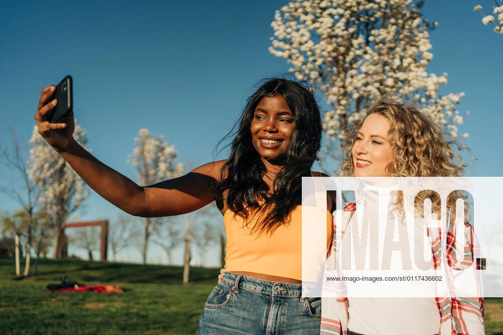 IMAGO Creative: N?chste Generation, Sozial- und klimabewusst Low angle of cheerful multiracial female best friends embracing in spring garden and taking self shot on smartphone on sunny day
