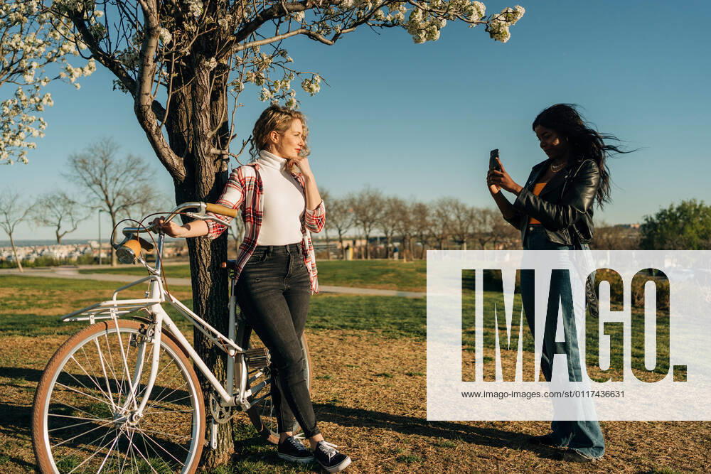 IMAGO Creative: Jahreszeit Fr?hling, Fr?hlingsanfang am 20. M?rz Side view of black woman taking photo of female friend standing with bicycle near tree in spring garden