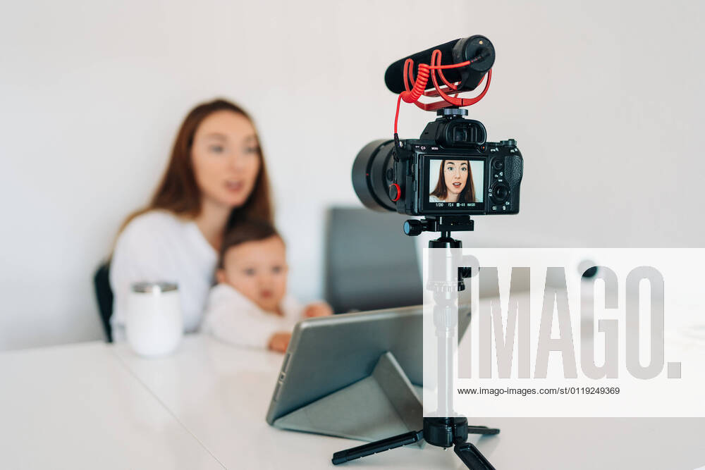 Blurred young mother with cute baby on laps talking and recording video on camera for personal blog while sitting at desk at home