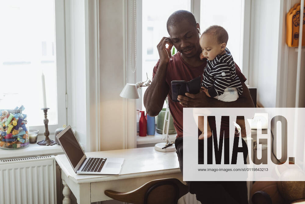 Male entrepreneur using smart phone while carrying baby boy at home office Sweden, Stockholm, model released, property released