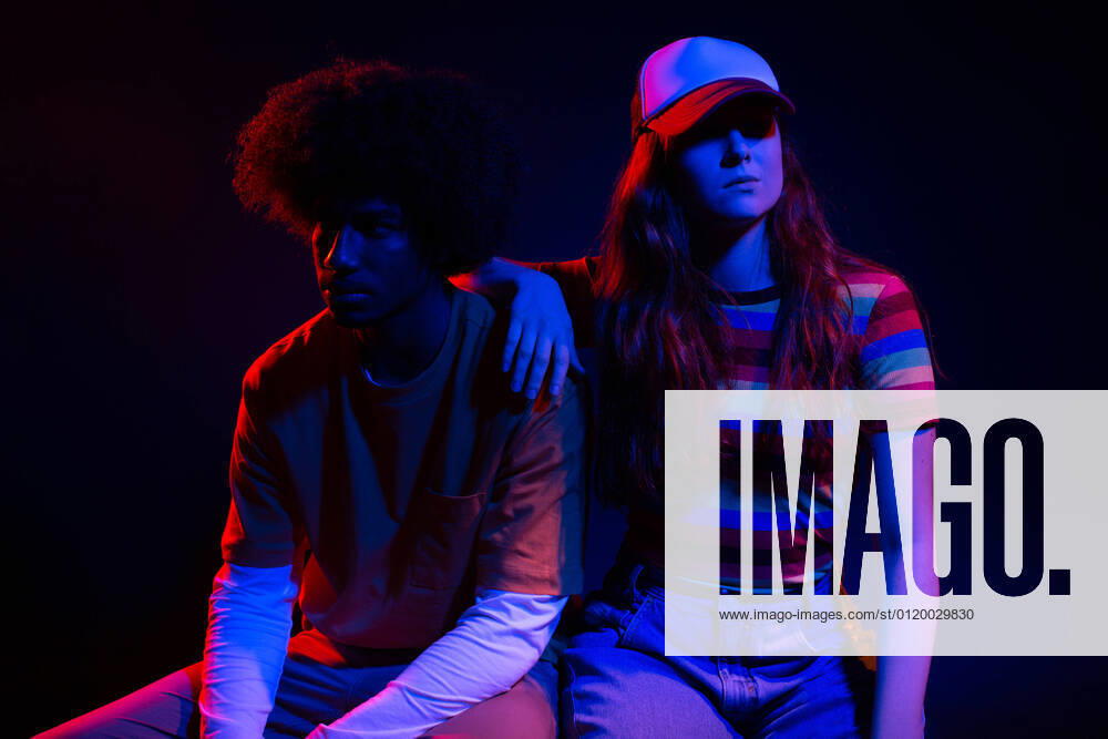 IMAGO Creative: N?chste Generation, Sozial- und klimabewusst Full body of self assured young female in trendy outfit sitting on chair with hand on shoulder of African American boyfriend in dark studio in neon lights, Model released