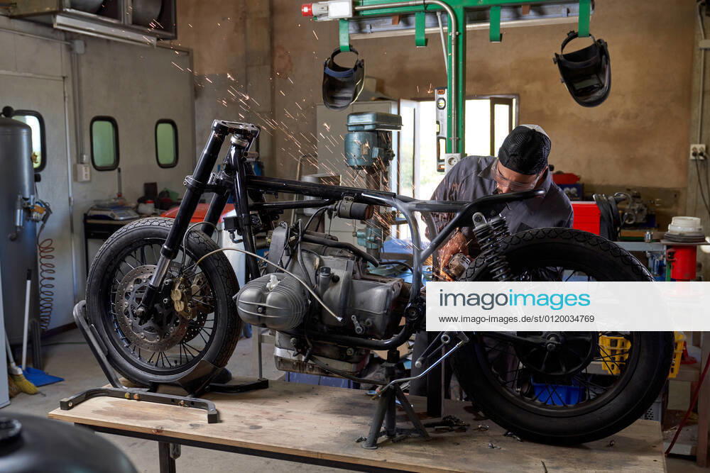Bearded worker in overall using angle grinder while polishing metal details of custom motorcycle in workshop