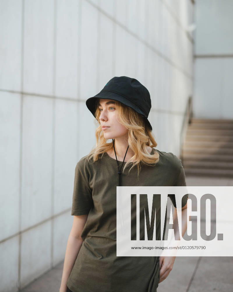 Young female in casual apparel looking away against concrete wall of modern building on urban pavement in daytime