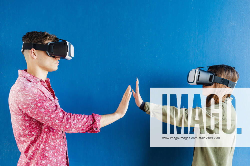 Friends with virtual reality headset stretching hands towards each other by blue wall model released Symbolfoto DLTSF01842